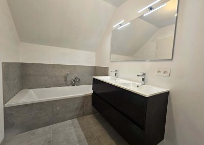 Salle de bain de la nouvelle construction Maison Braine-l'Alleud avec baignoire intégrée, double vasque moderne et carrelage gris.